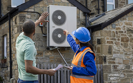 An air source heat pump on the side of a home being installed in the North East of England. The house is aiming to be sustainable. There is a construction worker with a hard hat and reflective jacket holding a digital tablet, they are both pointing to the heat pump.
